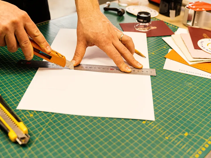 Hands trimming paper with a knife against a steel rule.